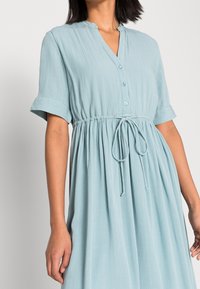 Ljusblå klänning i texturerad tyg, med v-ringning, korta uppvikta ärmar, knappstängning fram, justerbar dragsko i midjan och lös passform.