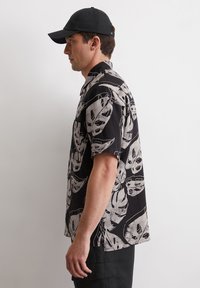 Schwarzes Kurzarmhemd mit weißem Blättermuster. Leichtes Material, mit Knopfleiste und klassischem Kragen. Getragen mit schwarzer Kappe und Hose.