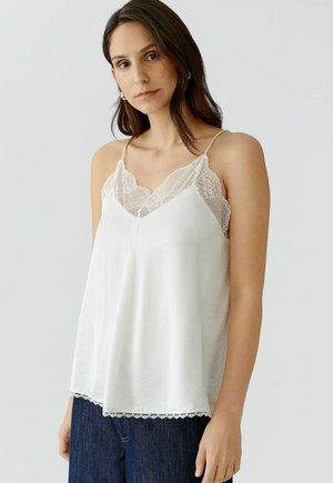 Junge Frau mit langen, dunklen Haaren trägt ein weißes, mit Spitze verziertes Camisole-Oberteil und dunkelblaue Jeans vor einem schlichten, hellen Hintergrund.