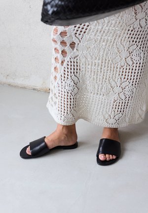 Füße in schwarzen Slide-Sandalen und einem weißen gehäkelten Rock, stehend auf einem hellgrauen Boden vor einer weißen Wand.