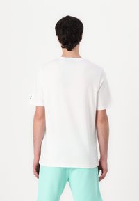 Camiseta branca de algodão com gola redonda e mangas curtas, vista de trás. Calções de alças verde-claro são usados por baixo.