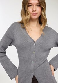 Cardigan grigio a coste con scollo a V, maniche lunghe e chiusura frontale con bottoni. Presenta bottoni colorati e un orlo posteriore più lungo.