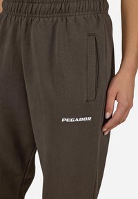 Brūnas sporta bikses ar elastīgu jostu un sānu kabatām. Uz kreisā kājas ir balta "PEGADOR" emblēma. Mīksta tekstūra, brīvs piegriezums.