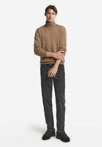 Ljusbrun polo-tröja med ribbad textur; kombinerad med mörkgrå slim-fit jeans och mörka skor. Modellen står med armarna i kors.