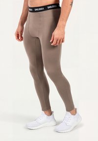 Mann trägt taupefarbene Kompressionsleggings mit einem schwarzen "SMILODOX"-Bund und weißen Sportschuhen, steht auf einem weißen Boden.