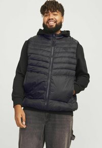 Polsterweste aus schwarzem Material mit Reißverschluss vorne, gestepptem Look und hohem Kragen, über einem schwarzen Hoodie getragen. Normale Passform, lässiges Design.