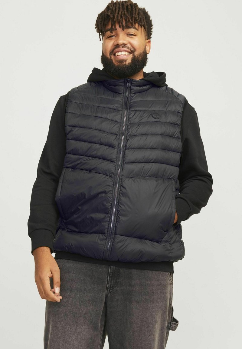 Polsterweste aus schwarzem Material mit Reißverschluss vorne, gestepptem Look und hohem Kragen, über einem schwarzen Hoodie getragen. Normale Passform, lässiges Design.