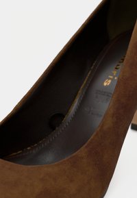 Καφέ σουέντ γόβα με στρογγυλεμένη μύτη, λεία εσωτερική επένδυση, μικρό τακούνι και διακριτικό ανάγλυφο λογότυπο στο πλάι. Υφή στην εξωτερική πλευρά.