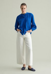 Camicia blu con bottoni e superficie testurizzata, pantaloni bianchi a gamba larga e mocassini navy a fantasia. Modello posa con le mani in tasca.