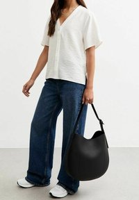 Černá kožená kabelka hobo s hladkou strukturou, širokým horním otevřením a minimalistickými designovými detaily, držená osobou v neformálním oblečení.