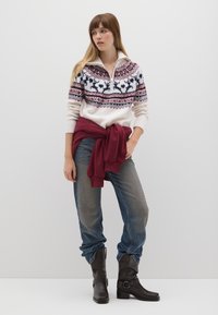 Maglione lavorato a maglia con base bianca, motivo rosso e blu, colletto con mezza zip; felpa maroon sovrapposta; jeans blu scuciti con accenti argentati; stivali marroni.