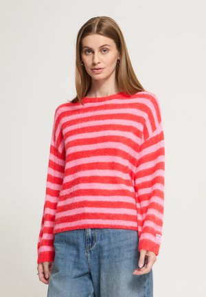 Giovane donna con capelli lisci castano chiaro che indossa un maglione peloso a righe rosa e rosse e jeans di denim blu su uno sfondo uniforme.
