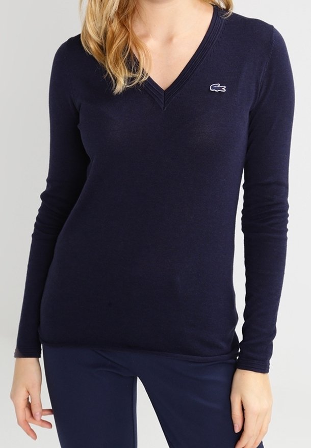 Femme portant un pull ajusté à col en V bleu marine à manches longues avec un petit logo crocodile sur le côté gauche de la poitrine.