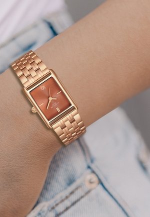 Montre rectangulaire en or rose avec un cadran en cuivre texturé. Dotée d'un bracelet en métal tissé et de marqueurs d'heure simples. Positionnée sur un poignet.