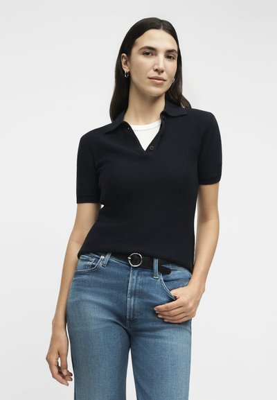 Γυναίκα με μακριά σκούρα μαλλιά, φορούσε μαύρο κοντομάνικο μπλουζάκι με γιακά πάνω από λευκό πουκάμισο, μπλε τζιν και μαύρη ζώνη, ποζάροντας μπροστά σε φωτεινό φόντο.