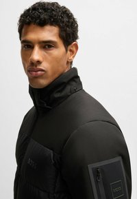 Man with curly hair wearing a black BOSS puffer jacket with high collar, visible logo on chest and zippered pocket on sleeve.