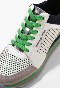 Baskets blanches avec lacets verts, bout en daim gris, perforations en forme d'étoile, logo « Michael More » et accents verts et bleus sur la semelle.