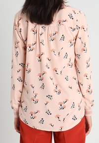 Blusa a maniche lunghe in morbido tessuto rosa con motivo floreale nei toni dell'arancio e del blu. Presenta una schiena arricciata e dettagli con polsini elastici.