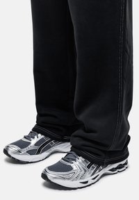 Scarpe sportive con una tomaia in rete argento e grigio, dettagli neri e suola bianca; indossate con pantaloni neri con cuciture bianche.