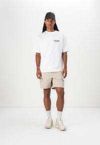 Weißes, oversized T-Shirt mit schwarzem Text, beigen Cargo-Shorts, grauen Socken und beigen Sneakers, ergänzt durch eine schwarze Mütze. Sauberes, minimalistisches Design.