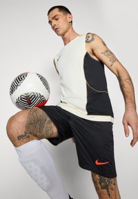 Nike brezrokavnik v beli in črni barvi z zlatimi detajli; črne kratke hlače; bele kolenske nogavice; črno-bež nogometna žoga z rdečimi detajli.