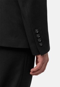 Černý saka s upraveným rukávem z texturované látky, se čtyřmi černými knoflíky s květinovými akcenty na manžetě a hladkým povrchem.