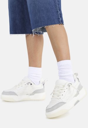 Witte en grijze sneakers met een gestructureerde afwerking, voorzien van een dikke zool, veters en demping. Gecombineerd met witte geribde sokken en denim shorts.
