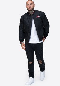 Czarna bomber jacket z czerwonymi i białymi prążkowanymi mankietami, zestawiona z przetartymi czarnymi dżinsami i białymi sneakersami.