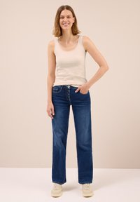 Donna sorridente in piedi con una mano in tasca, indossa una canotta color crema, jeans blu scuro e scarpe da ginnastica beige, sullo sfondo semplice.