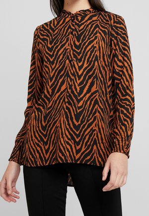 Damenbluse mit langen Ärmeln und hohem Kragen mit schwarz-braunem Tigerstreifenmuster, getragen zu schwarzen Hosen.