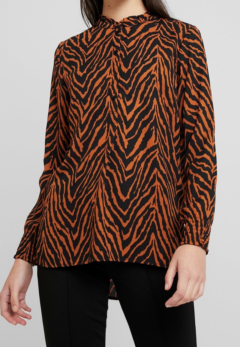 Blouse femme à manches longues avec col montant et motif rayures de tigre noir et marron, portée avec un pantalon noir.