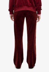 Person iført bourgogne velour bukser og matchende sweatshirt, stående med afslappede arme langs siderne, vist fra ryggen mod hvid baggrund.