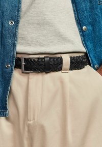Sort vævet læderbælte med en rektangulær sølvspænde, båret over lyse beige bukser og en grå top, der er stukket ind under en blå denimskjorte.