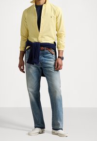 Man i en gul knäppt skjorta, blå jeans, vita sneakers, en mörk tröja knuten i midjan, brun bälte och en armbandsklocka.