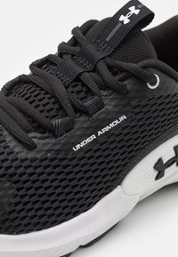 Zwarte sportieve schoen met een textuur van mesh bovenwerk, voorzien van een gladde zwarte materiaal en witte accenten. "UNDER ARMOUR" logo aan de zijkant.