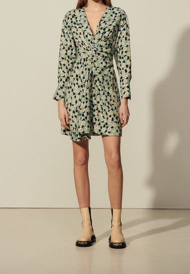 Femme portant une mini-robe verte à motifs floraux, manches longues, col en V profond et bottines beiges, debout devant un fond uni.