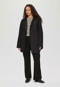 Manteau trench noir à double boutonnage avec un large col, porté sur un haut rayé noir et blanc et un pantalon large noir.