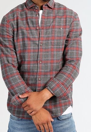 Hombre con camisa de cuadros roja y gris abotonada sobre camiseta blanca, con los brazos cruzados y vaqueros azules.