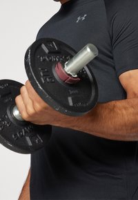 Svart viktdumbell som hålls i en hand, med ett metallhandtag och gummiklädda ändar. Bakgrunden är neutral med en åtsittande svart tröja synlig.