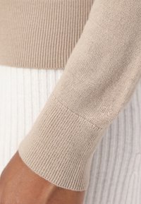 Bežový pletený svetr s rukávem s žebrovanou strukturou, obsahující jemný detail loga a kombinovaný s bílým žebrovaným oděvem pod ním.