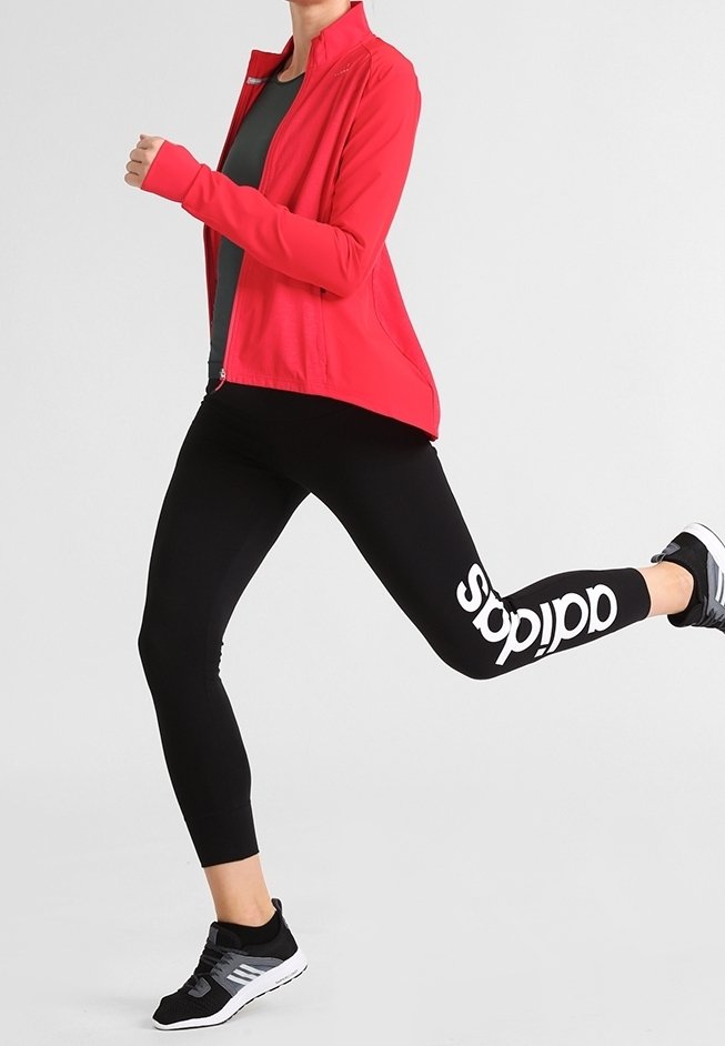 Personne en leggings Adidas noirs et chaussures de course noires en train de jogger, portant une veste rouge et un t-shirt gris foncé.