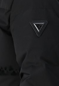 Svart tyg med en sydd triangel-formad Guess-logotyp i ett metalliskt silverfärgat kantband och graverad text på jackärmen.