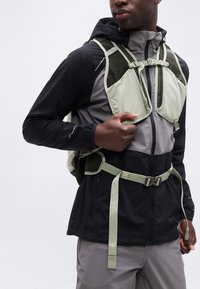 Άνδρας που φοράει μαύρο και γκρι εξωτερικό μπουφάν και ανοιχτοπράσινο γιλέκο πεζοπορίας με ρυθμιζόμενους ιμάντες σε απλό φόντο.