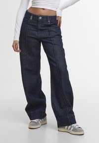 Jean bleu foncé à jambes larges porté avec des baskets grises et blanches et un crop top blanc à manches longues, main posée sur la hanche, debout sur un sol blanc.