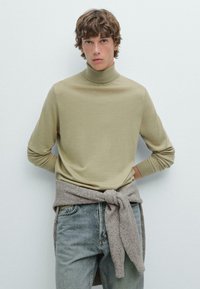 Ljus olivgrön polotröja i mjukt tyg, kombinerad med lösa, bleknade blå jeans. En gråbrun stickad tröja är knuten runt midjan.