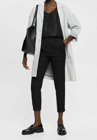 Šedý dlouhý kabát přes černý strukturovaný top, černé kalhoty a lesklé černé boty. Černá taška přes rameno, jednoduchý stříbrný náhrdelník a ohrnuté rukávy.