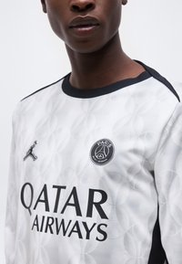 Κοντινό πλάνο ενός ατόμου που φοράει λευκή φανέλα ποδοσφαίρου της Παρί Σεν Ζερμέν με μαύρο γιακά και το λογότυπο της Qatar Airways.