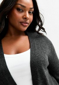 Chandail cardigan gris foncé en matériau doux, présentant un profond décolleté en V. Superposé sur un débardeur à côtes blanc. Texture lisse.