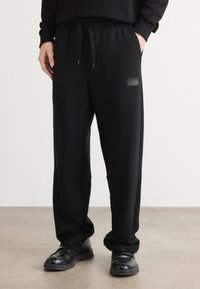 Zwarte stretchstof joggers met een elastische tailleband en trekkoord, zijzakken en een klein logopatch op de dij.