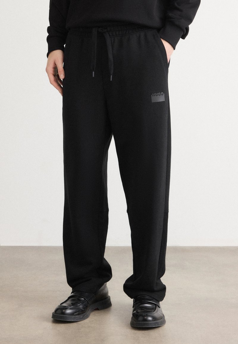 Zwarte stretchstof joggers met een elastische tailleband en trekkoord, zijzakken en een klein logopatch op de dij.